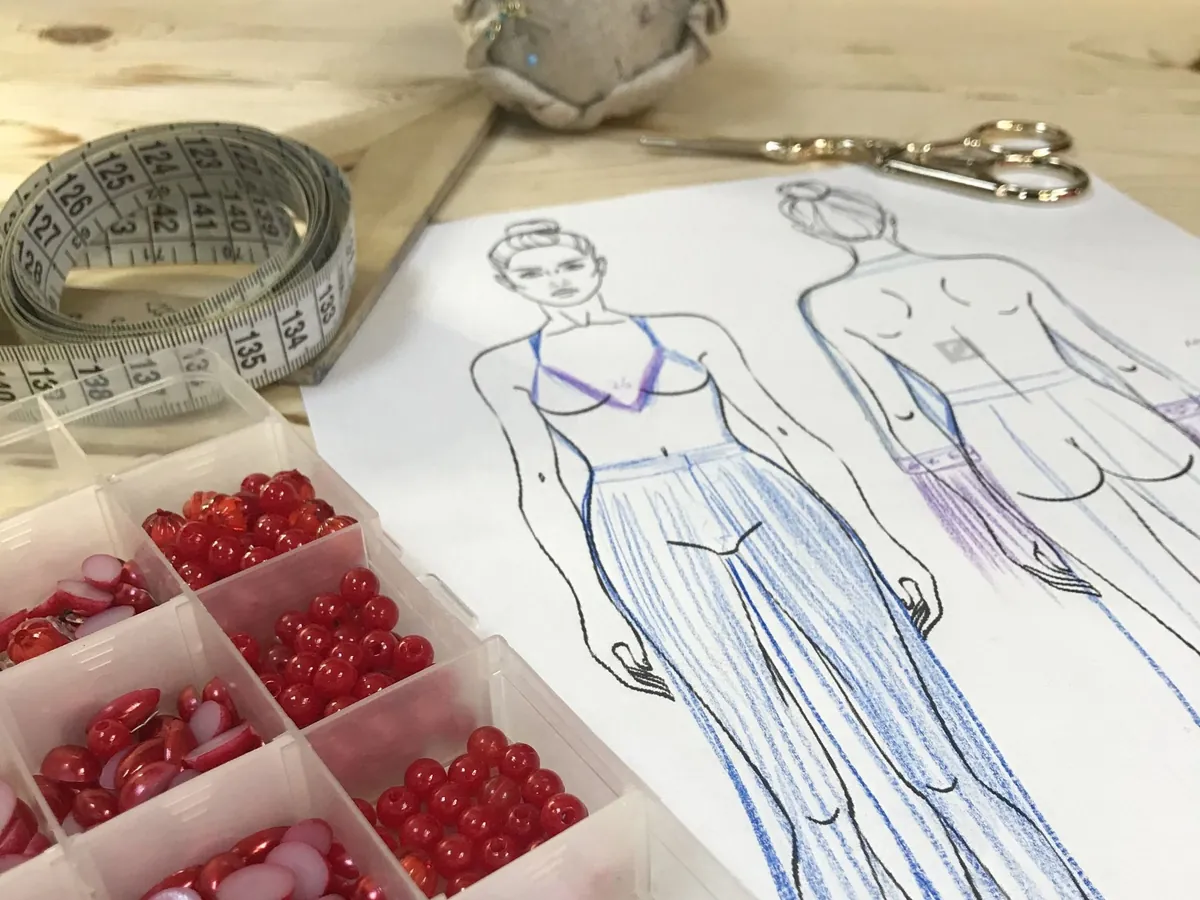 Figurines dibujados a lápiz y coloreados en azul, junto a una cinta métrica, un alfiletero, una tijera y una caja de botones rojos